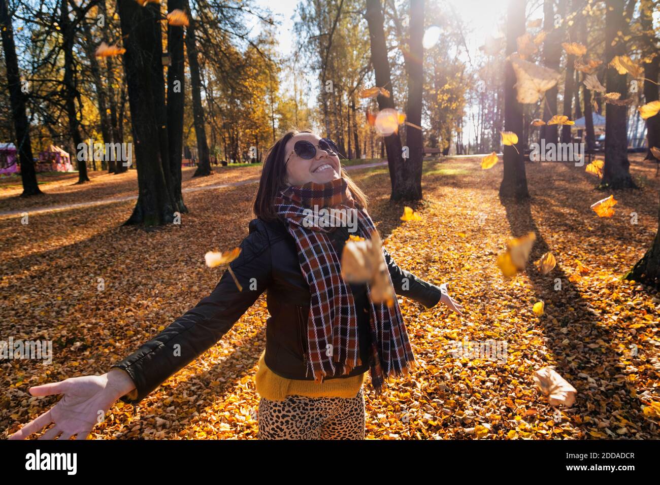 Sorridente bella donna con le braccia allungate godendosi mentre si sta sotto cadere foglie secche al parco durante l'autunno Foto Stock