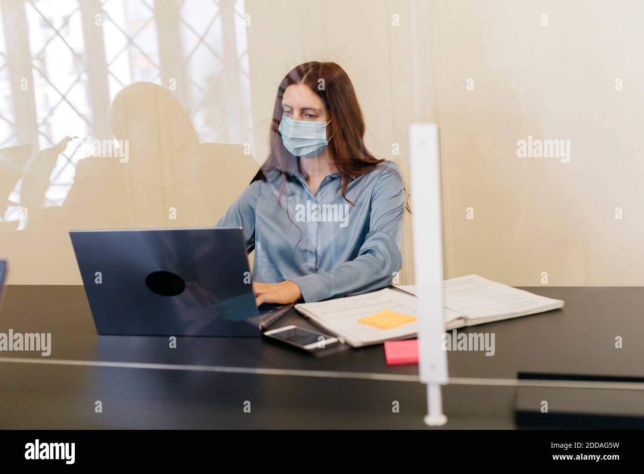 Donna imprenditore con maschera protettiva che lavora sul laptop sul luogo di lavoro vista attraverso il vetro del finestrino Foto Stock