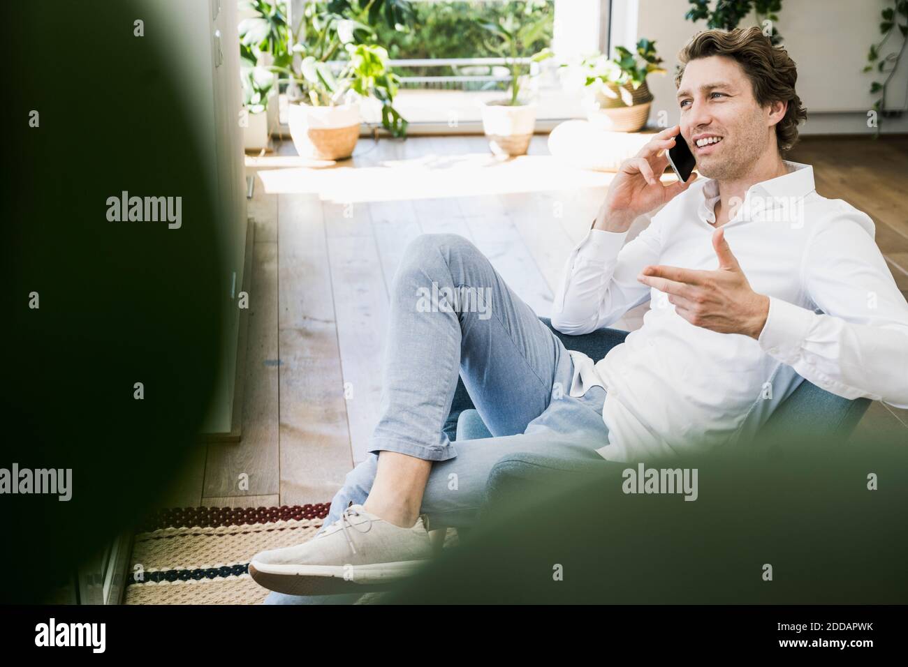 Medio adulto che usa lo smartphone mentre si siede sulla sedia a. casa Foto Stock