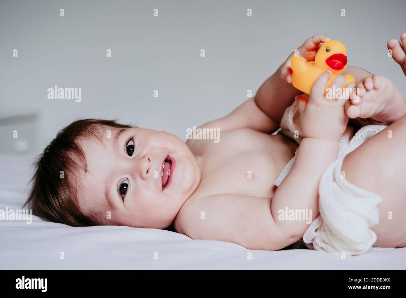 Sorridente simpatico bambino che gioca con il giocattolo d'anatra mentre si sdraiava giù sul letto a casa Foto Stock