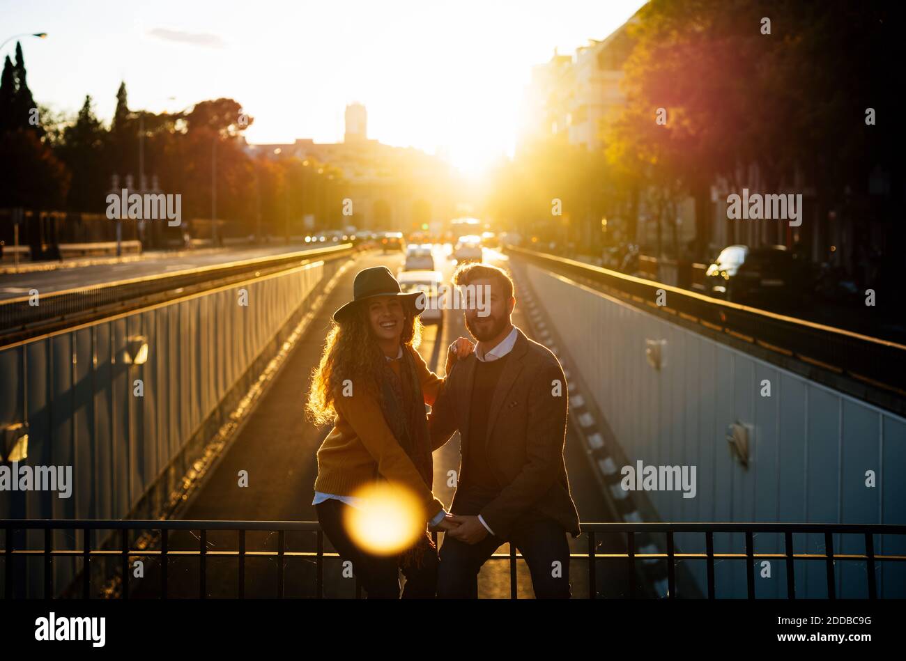 Coppia che tiene le mani mentre si siede sulla ringhiera del ponte insieme a. città durante il tramonto Foto Stock