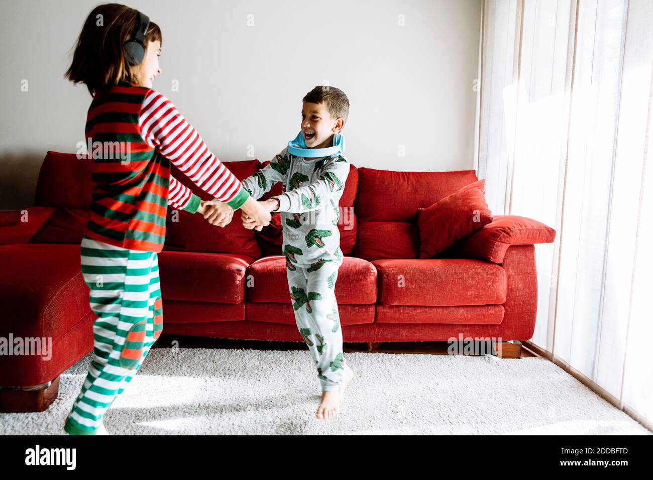 Fratello e sorella con le cuffie che tengono le mani mentre ballano contro divano in soggiorno Foto Stock