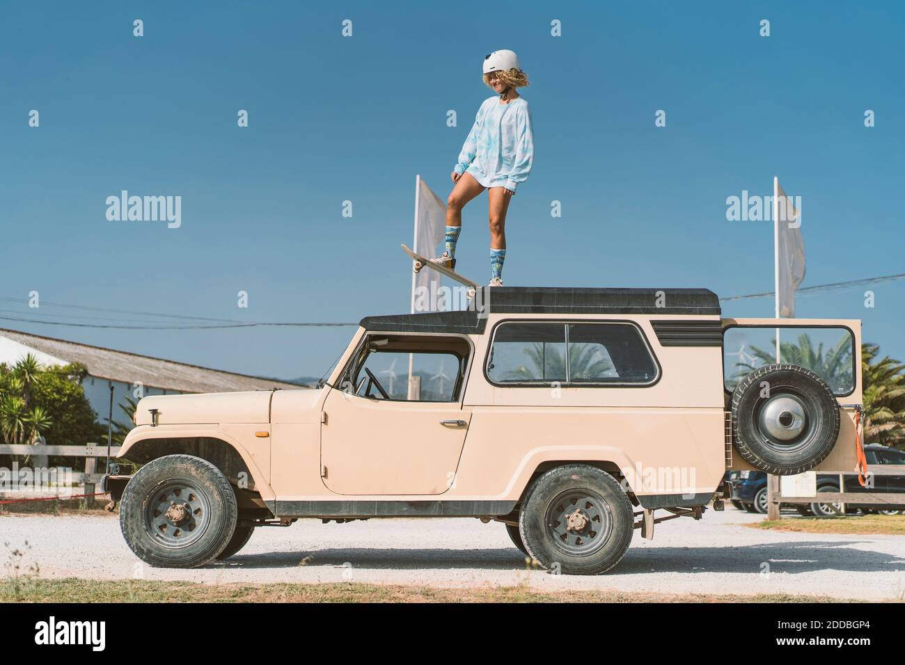 Giovane donna che indossa un casco in piedi con skateboard su un vecchio fuoristrada veicolo contro il cielo blu durante la giornata di sole Foto Stock