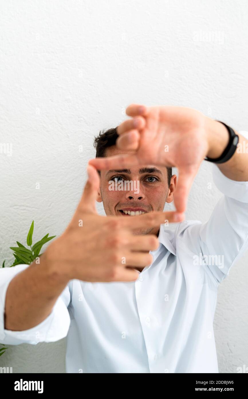 Uomo d'affari sorridente che fa la forma quadrata dalle dita e dai pollici contro parete bianca in ufficio Foto Stock