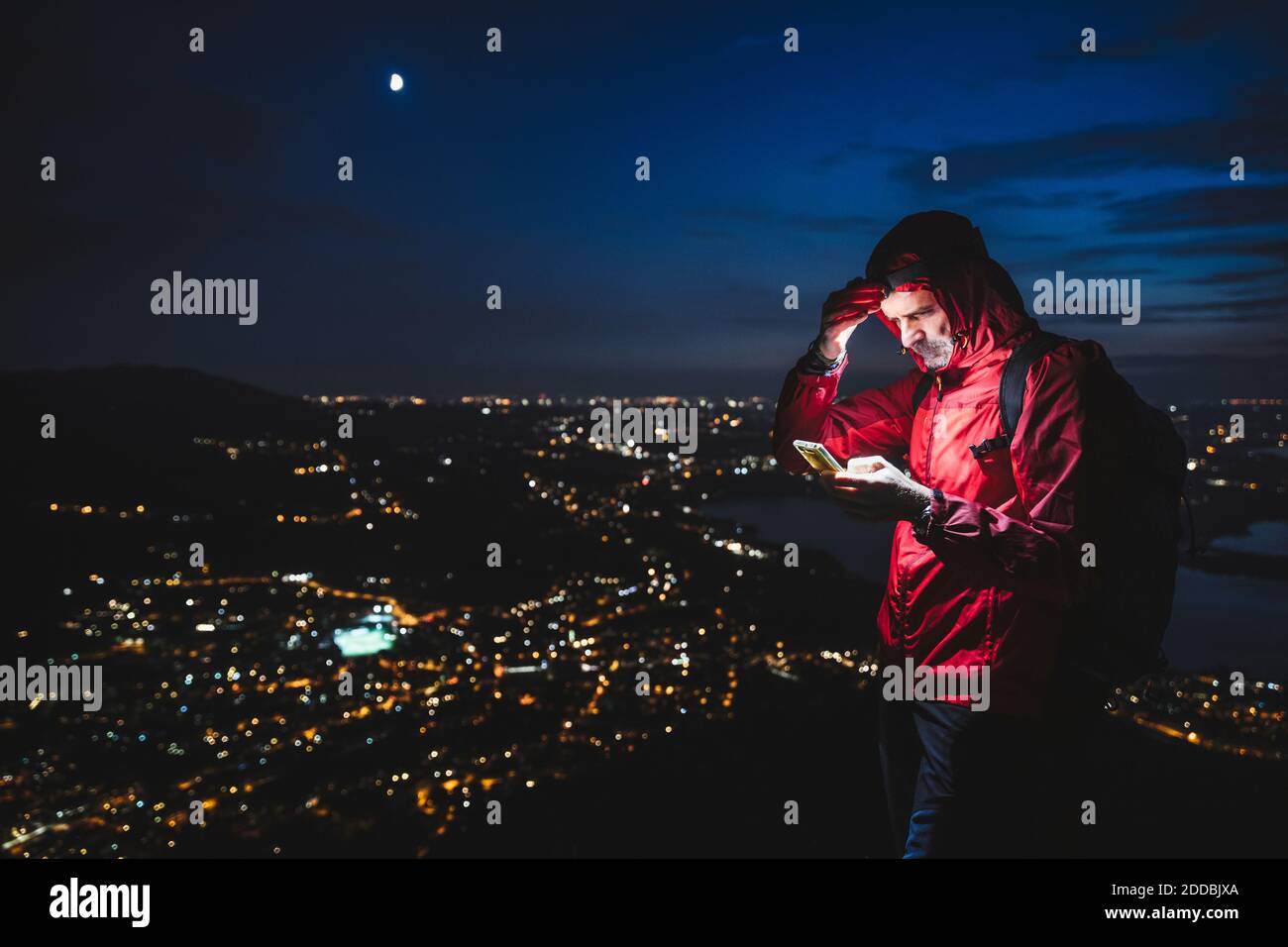 Escursionista maschile maturo utilizzando smartphone con città illuminata contro cielo di notte Foto Stock