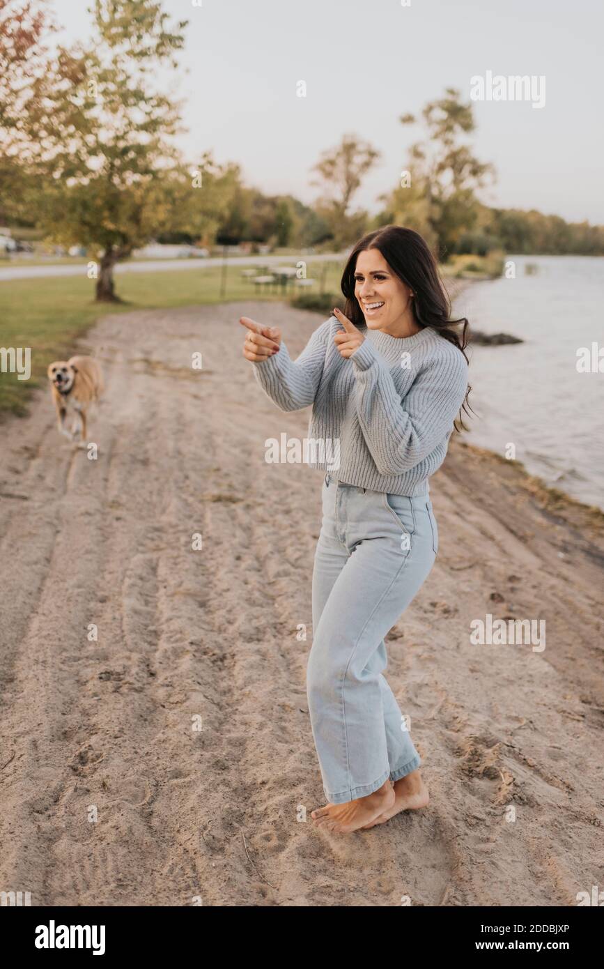 Donna sorridente che danzano mentre si sta con il cane che corre in background in riva al lago Foto Stock