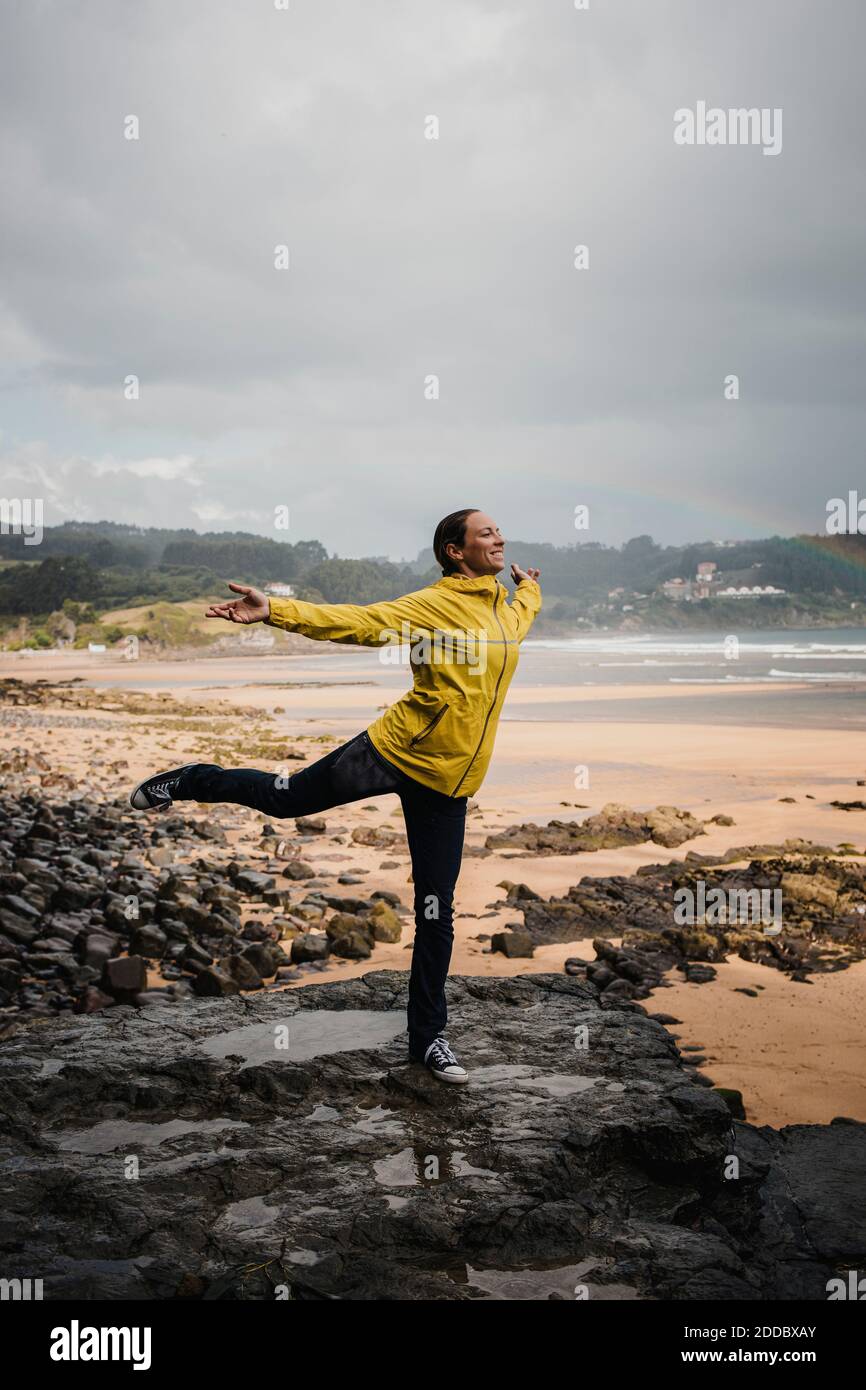 Giocosa donna in impermeabile in piedi su una gamba a. spiaggia durante la stagione delle piogge Foto Stock