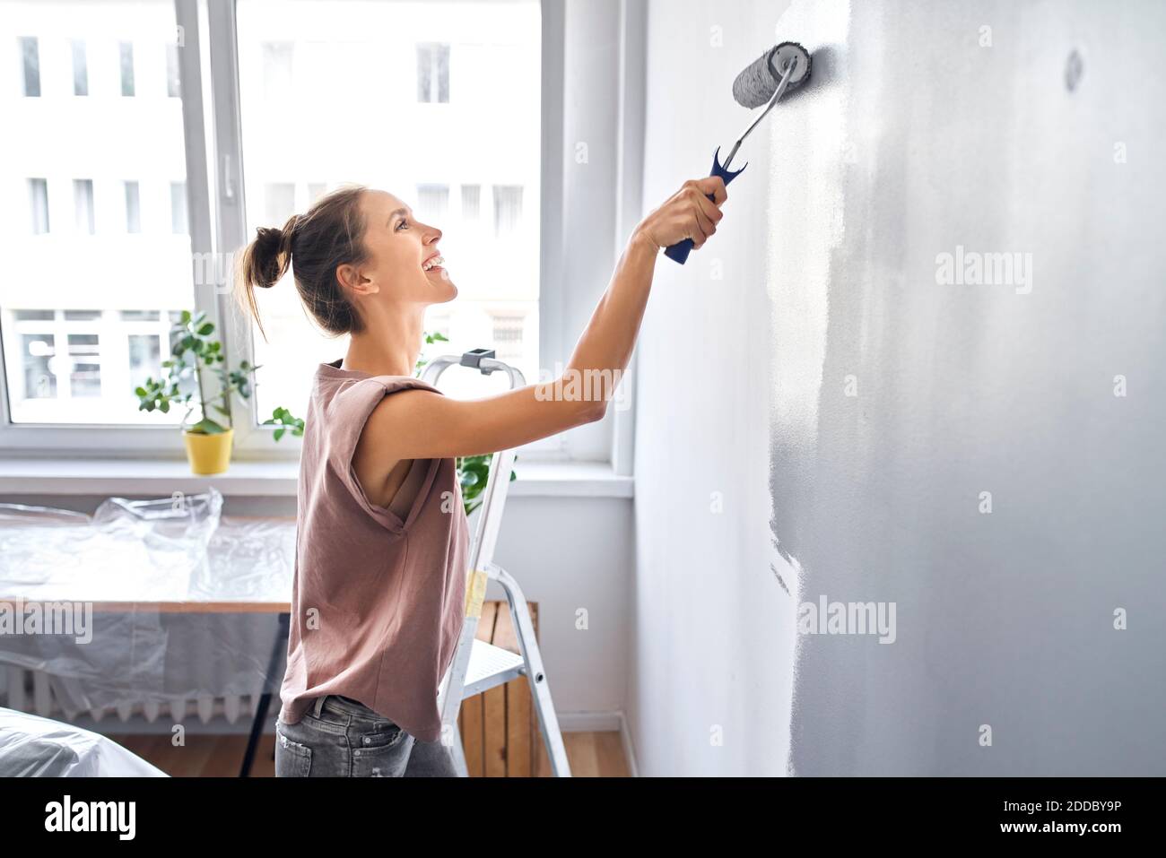 Donna sorridente pittura muro con rullo di vernice mentre in piedi a. casa Foto Stock