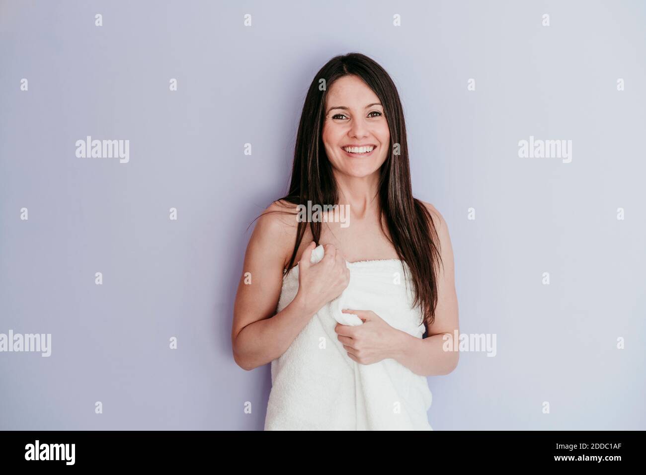 Sorridente bella donna avvolta in asciugamano in piedi contro parete a. casa Foto Stock