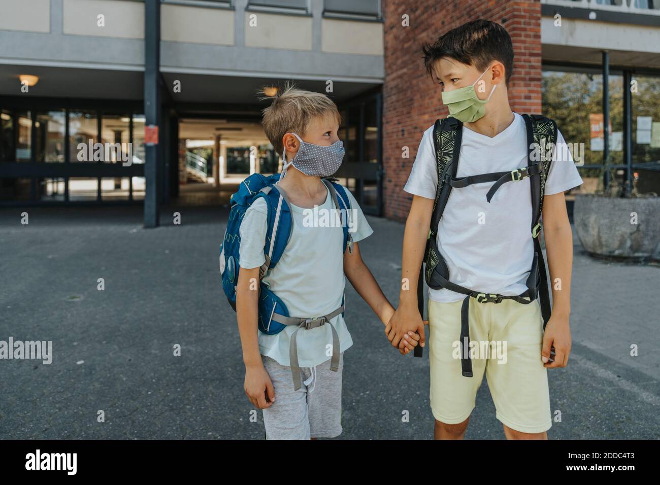 I fratelli indossano una maschera protettiva che tiene le mani in piedi sulla strada di fronte all'edificio scolastico Foto Stock