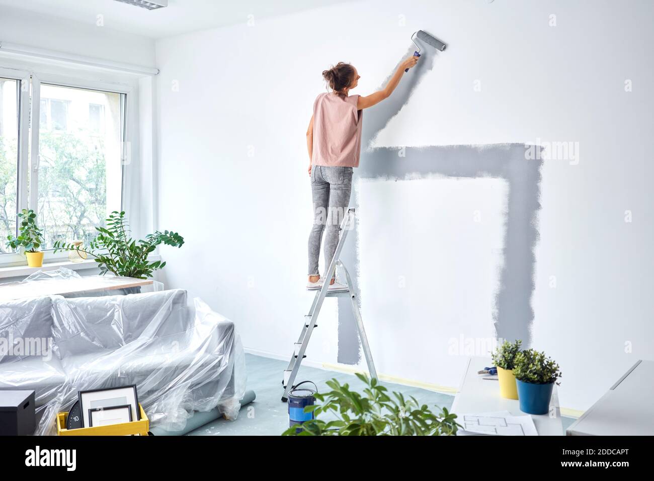 Giovane donna pittura muro con rullo dolore mentre si sta in piedi scala a casa Foto Stock