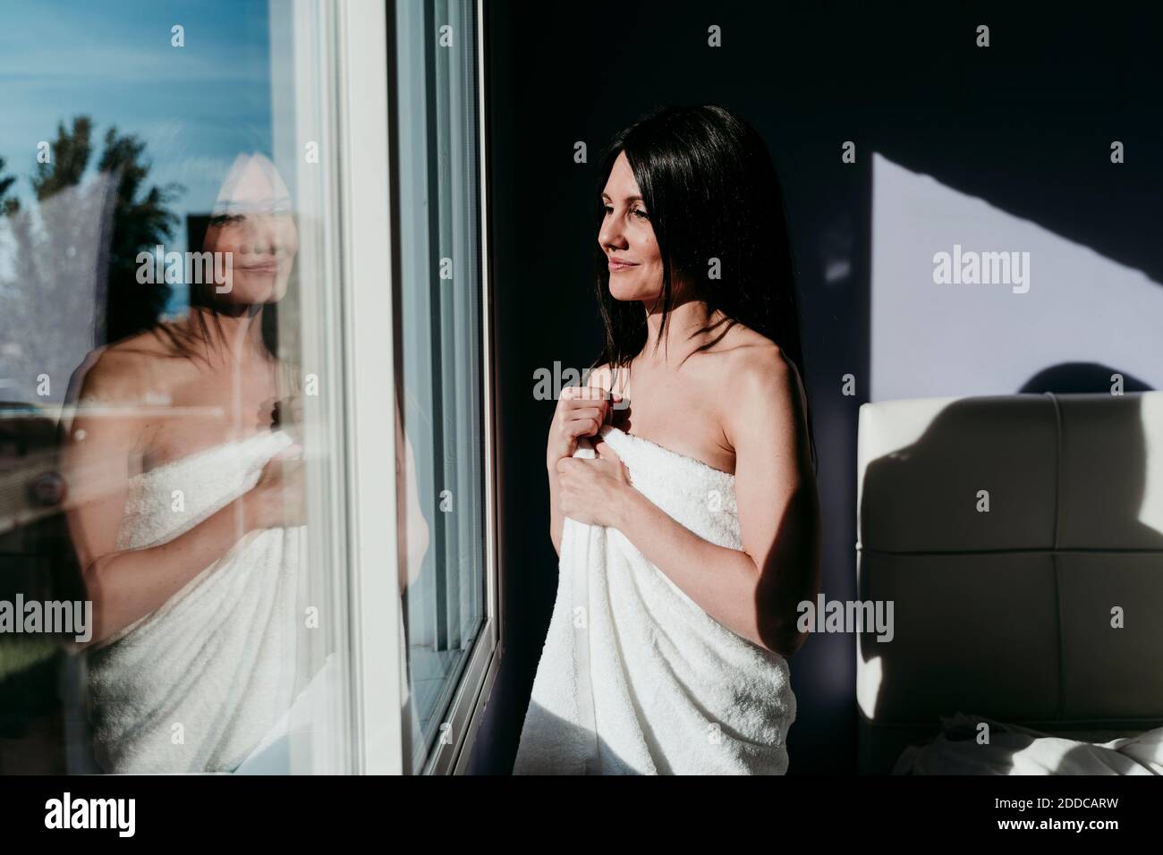 Donna sorridente e premurosa avvolta in asciugamano in piedi dalla finestra a. casa Foto Stock