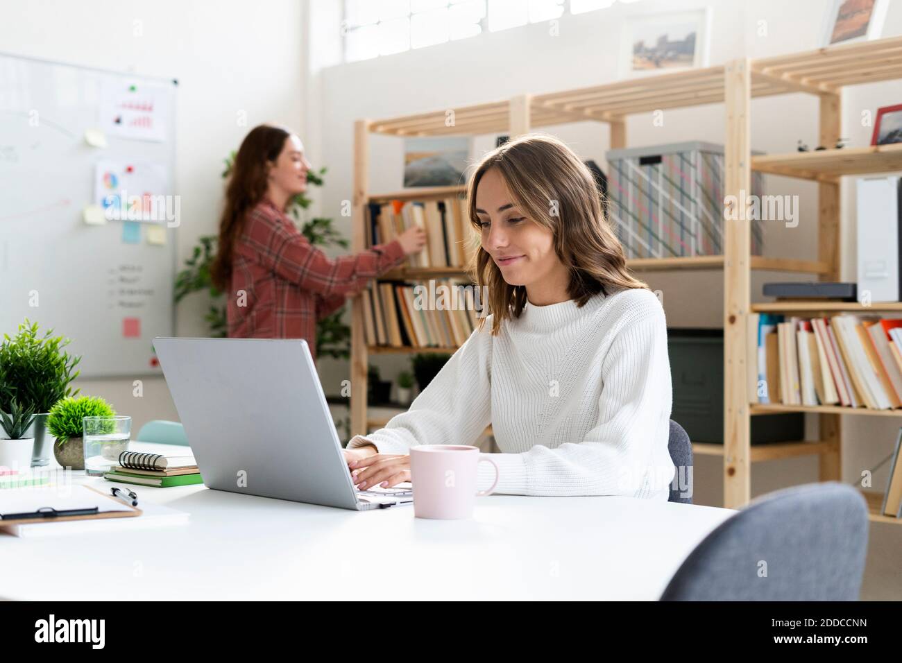 Donna d'affari sorridente che lavora su un computer portatile con un collega in piedi in background in ufficio Foto Stock
