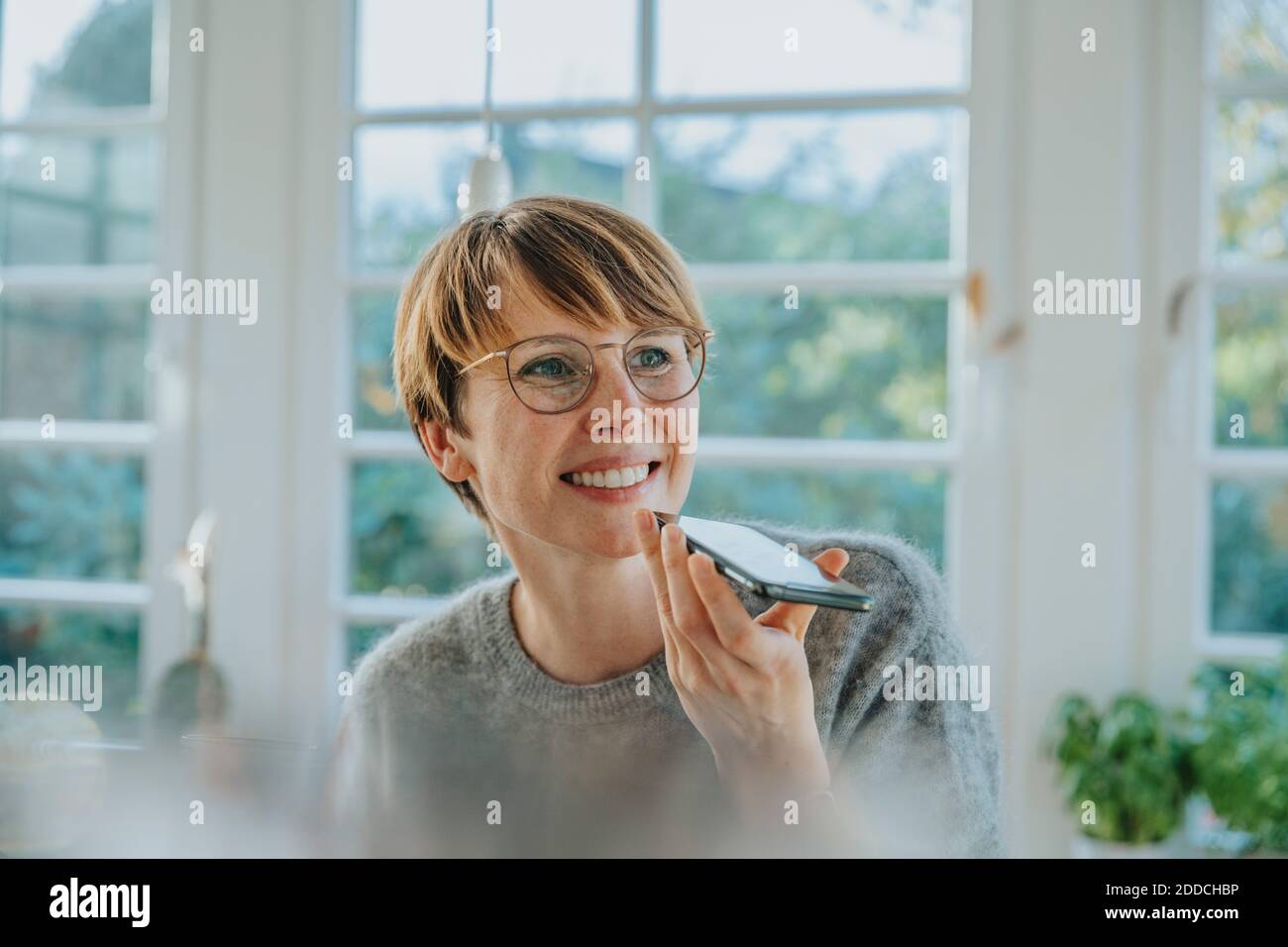 Felice donna mid adulta che invia messaggi vocali sullo smartphone mentre stare a casa Foto Stock