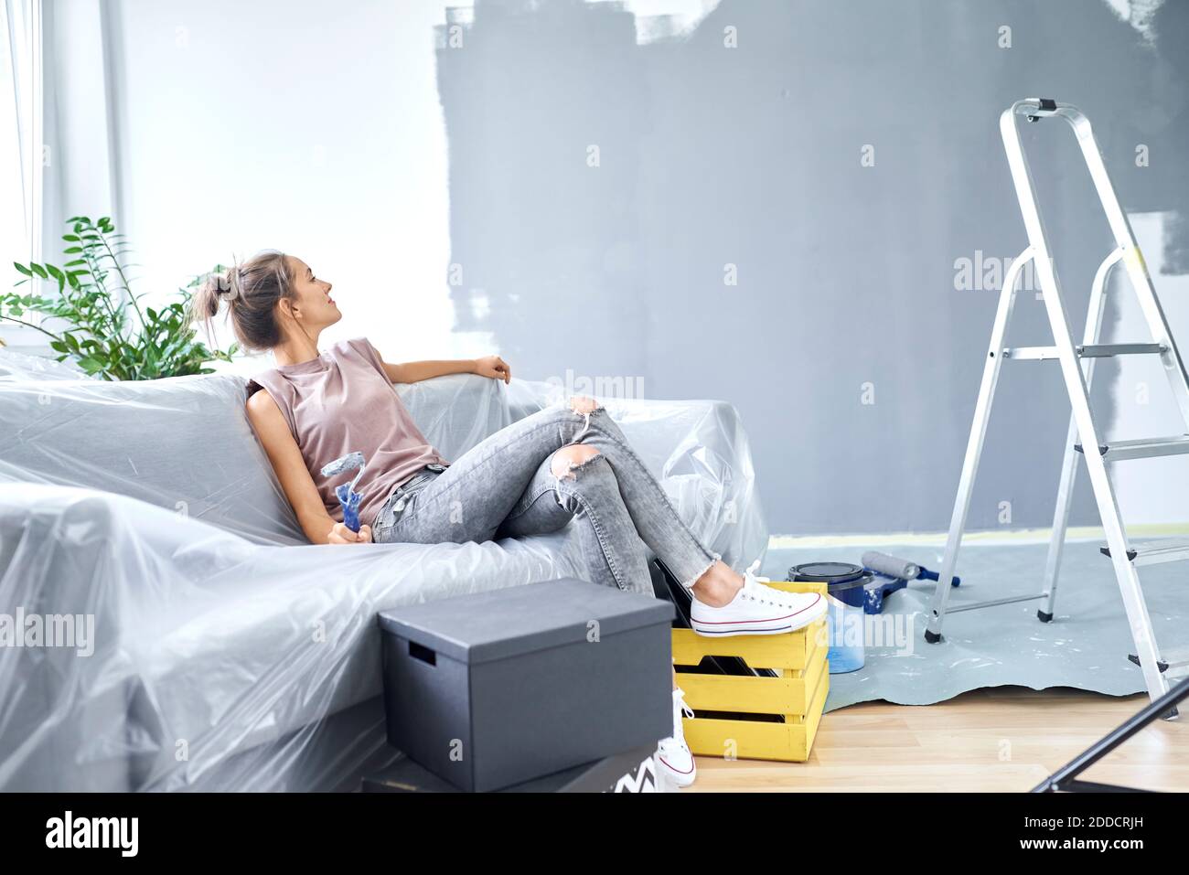 Donna con rullo di vernice che guarda il muro mentre si siede divano a casa Foto Stock