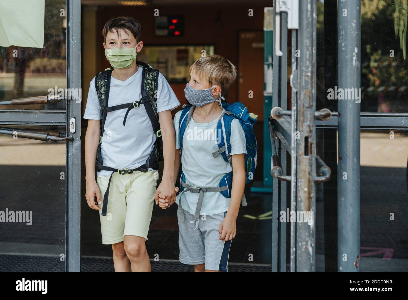 I fratelli indossano una maschera protettiva che tiene le mani che escono edificio scolastico Foto Stock