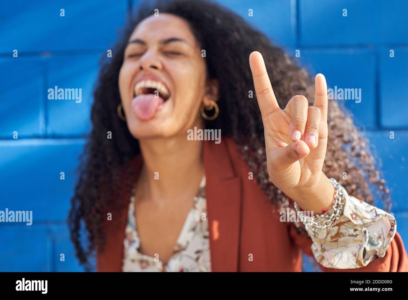 Giovane donna che si stacca la lingua facendo il segno del corno contro il blu parete Foto Stock
