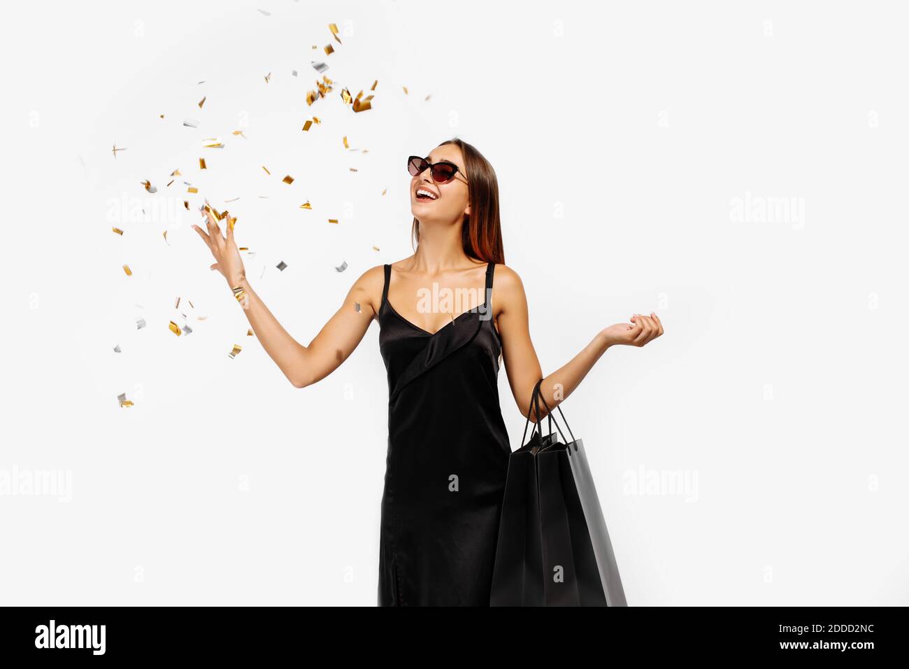 Giovane donna entusiasta, in abito nero, con borse, festeggiamenti e lancio di confetti su sfondo bianco, concept, shopping, Black Friday Foto Stock