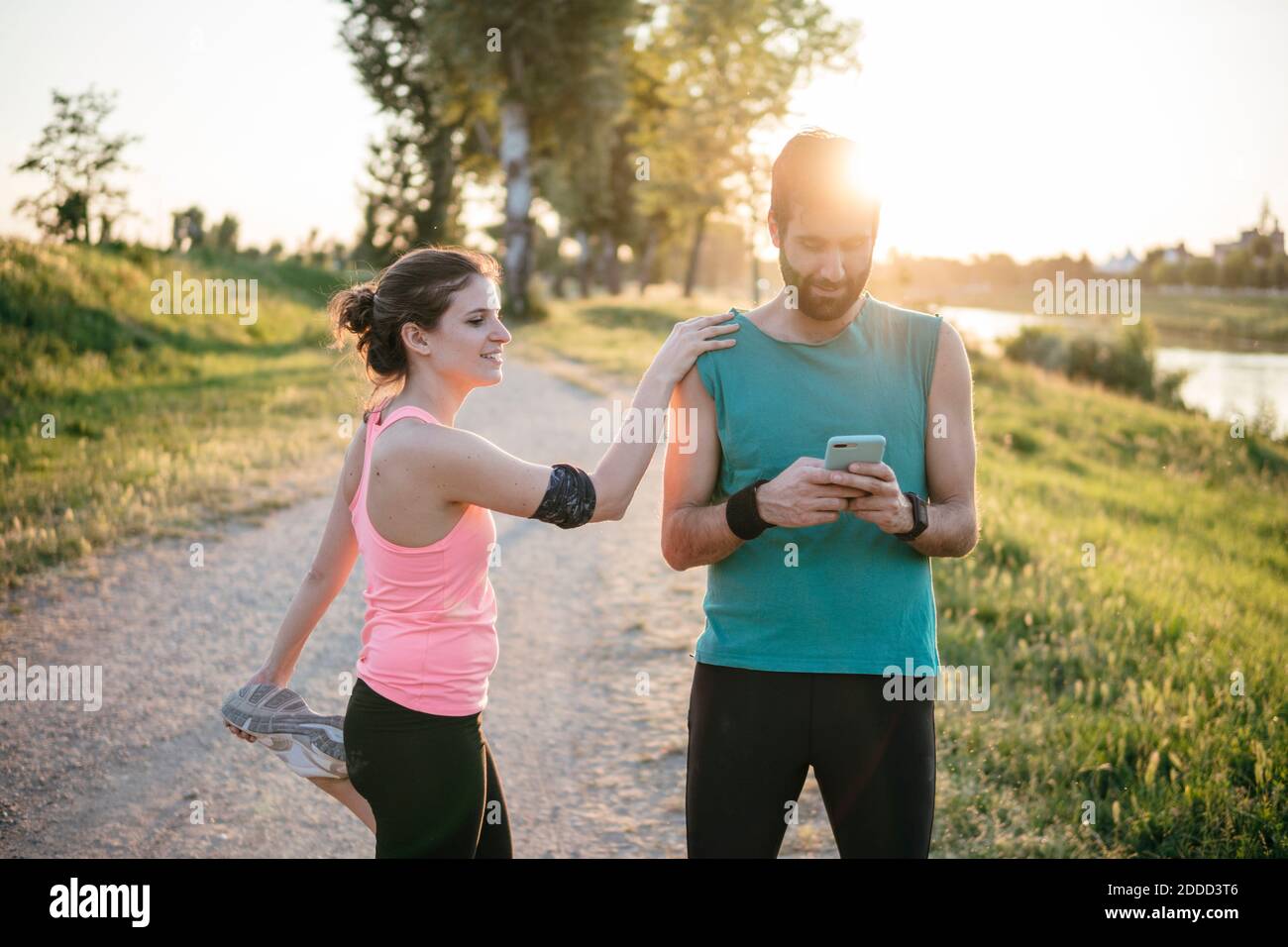 Atleta maschile che usa lo smartphone mentre la donna sportiva con la gamba in su in piedi nel parco durante il tramonto Foto Stock
