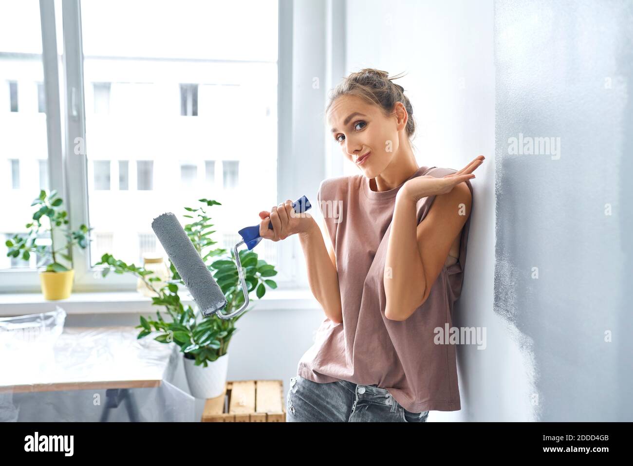 Bella giovane donna che tiene rullo di vernice mentre si appoggia sulla parete a casa Foto Stock