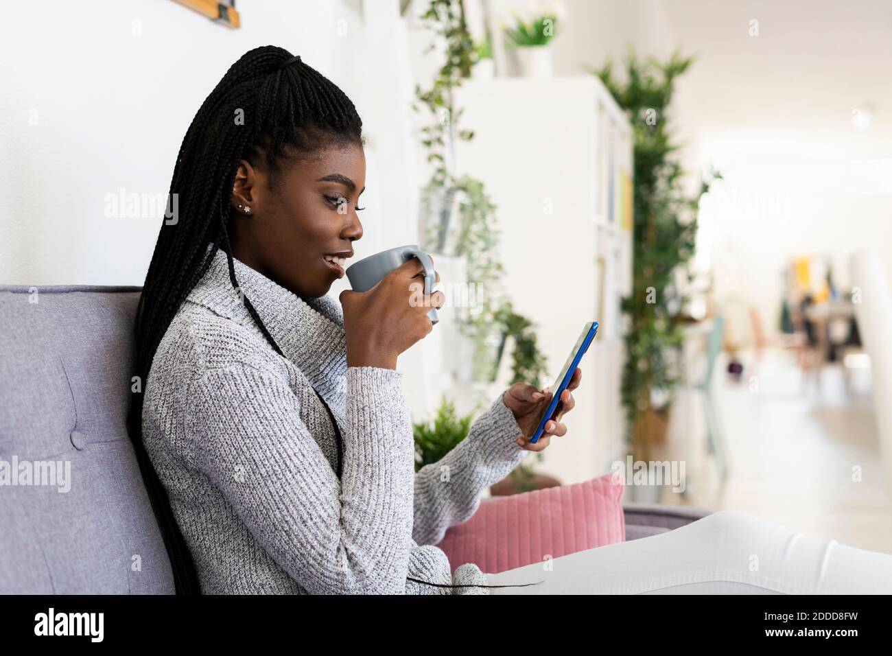 Giovane donna che beve caffè mentre invia messaggi di testo sullo smartphone a casa Foto Stock