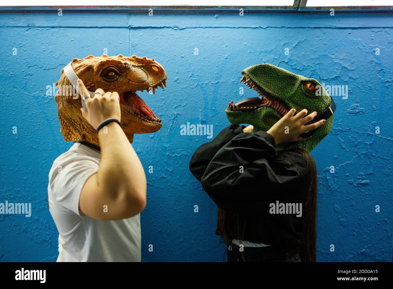 Amici di sesso maschile e femminile che ascoltano la musica con le cuffie mentre indossare una maschera di dinosauro contro la parete blu Foto Stock