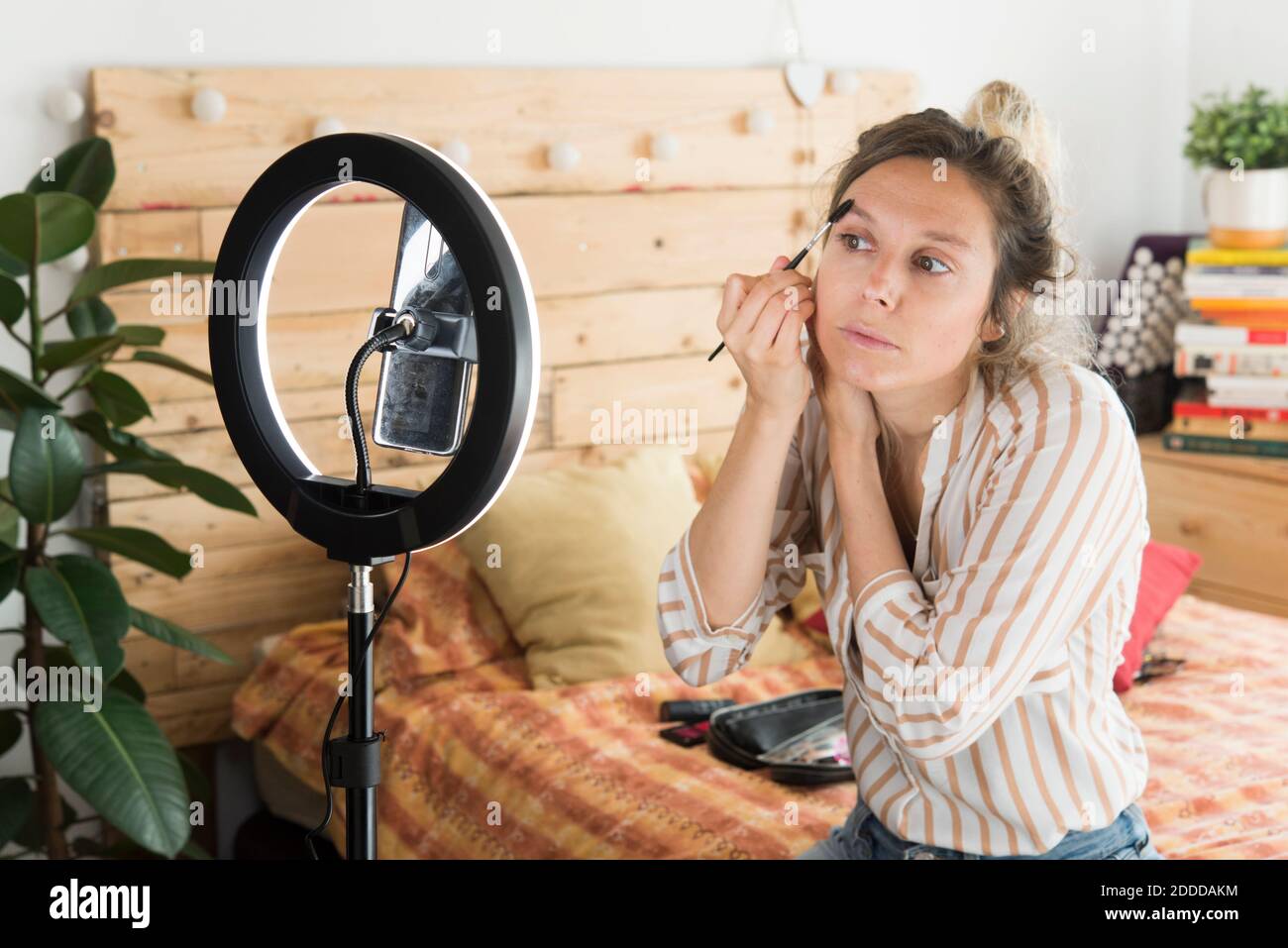 Donna influencer spazzolando le sopracciglia mentre vlogging attraverso smartphone seduto a casa Foto Stock