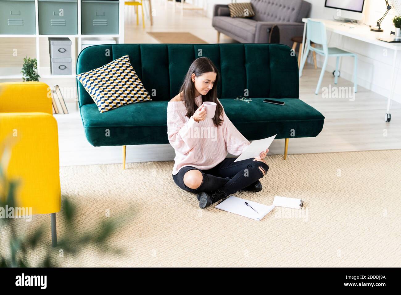 Bella giovane donna che beve il caffè mentre legge la carta per nota cuscini sul tappeto contro il divano a casa Foto Stock