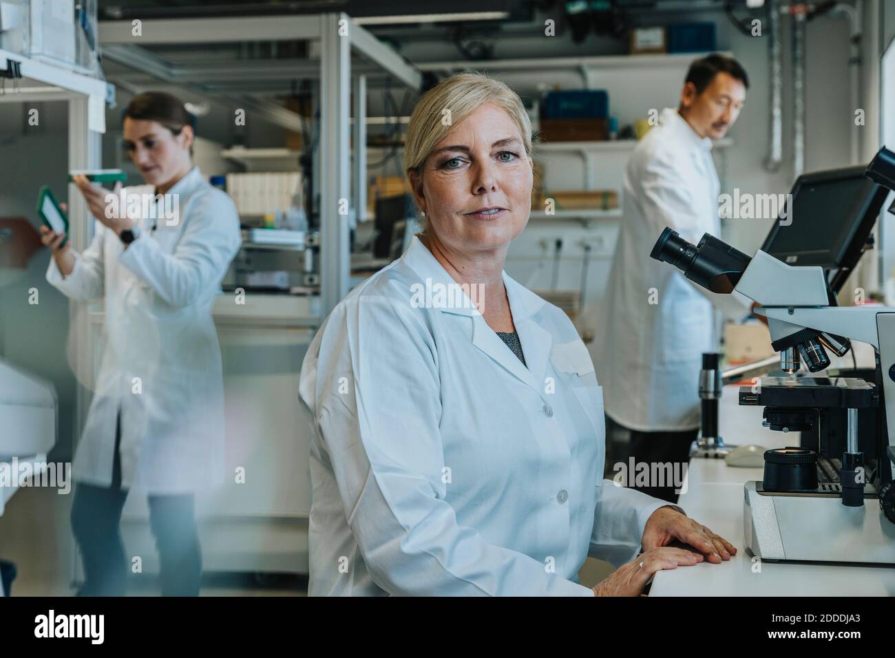 Donna scienziata seduta al microscopio con collaboratore che lavora in background in laboratorio Foto Stock
