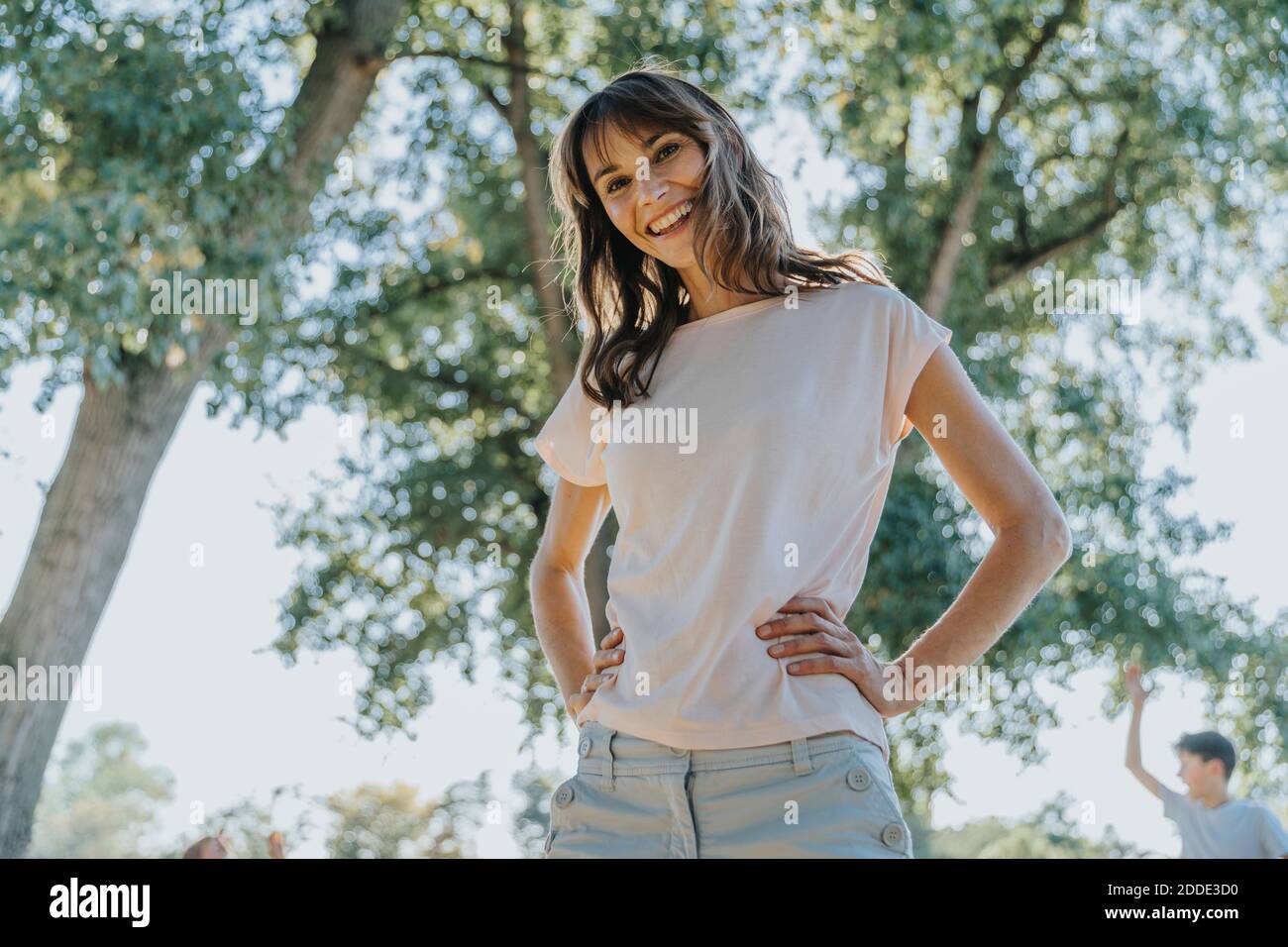 Donna matura sorridente con mano sull'anca in piedi in pubblico parcheggia durante la giornata di sole Foto Stock