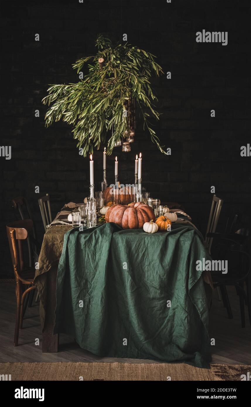 Elegante ambiente rustico per il giorno del Ringraziamento. Tavolo pieno con tovaglia di lino verde, stoviglie, bicchieri e posate decorate con zucche, portacandele in vetro e rami di ulivo Foto Stock