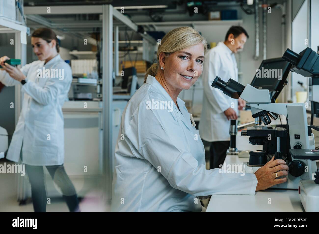 Scienziato sorridente seduto al microscopio con colleghi che lavorano in background in laboratorio Foto Stock