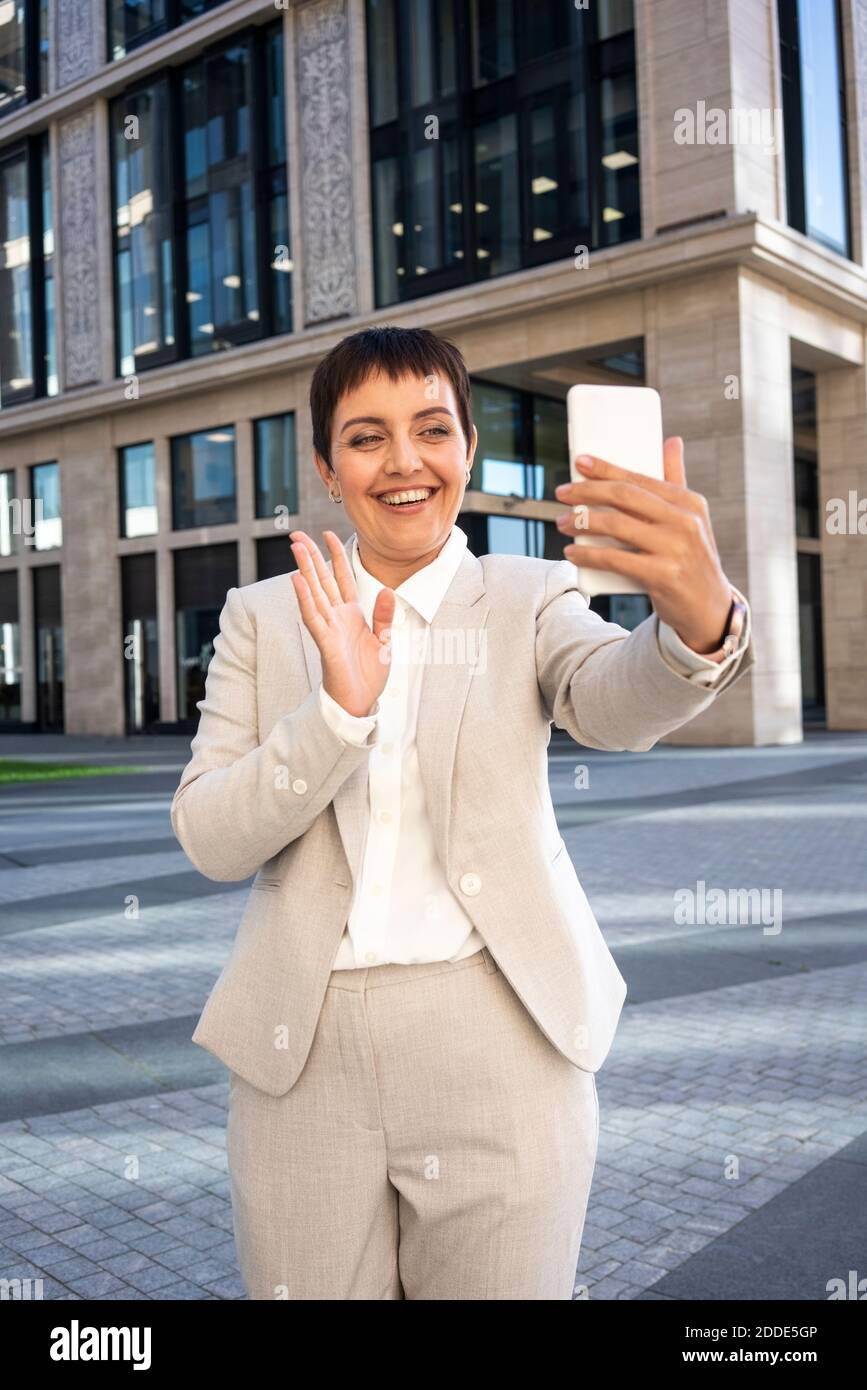 Donna che agita la mano per videochiamare sul telefono cellulare mentre in piedi contro un moderno edificio di uffici Foto Stock