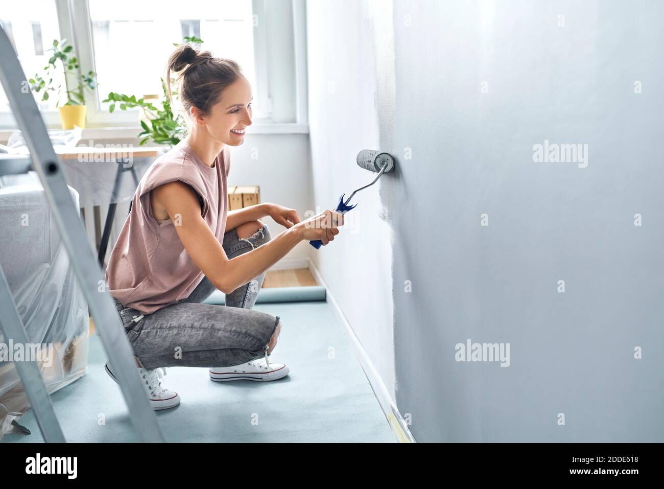 Giovane donna pittura muro con rullo di vernice mentre accovacciando a. casa Foto Stock