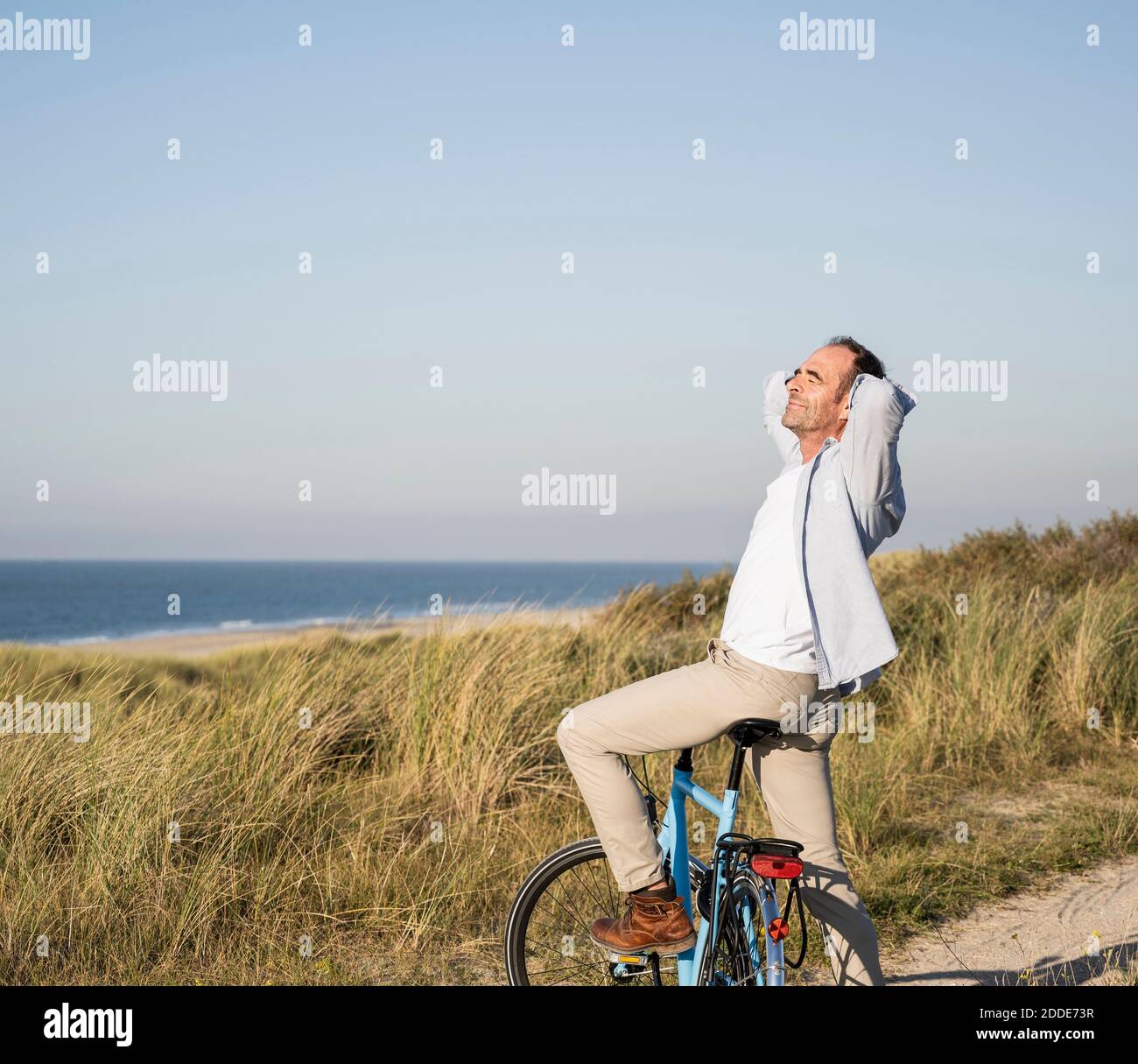 Uomo maturo in bicicletta con le mani dietro la testa in spiaggia contro il cielo limpido Foto Stock