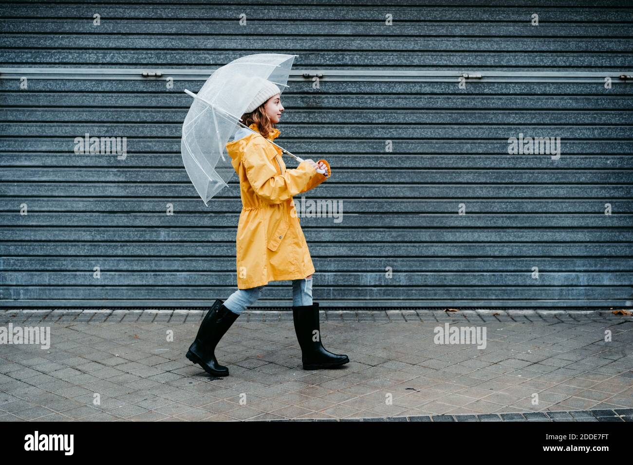 Ragazza che indossa impermeabile e scarpone da salto che tiene l'ombrello mentre cammina sul marciapiede Foto Stock