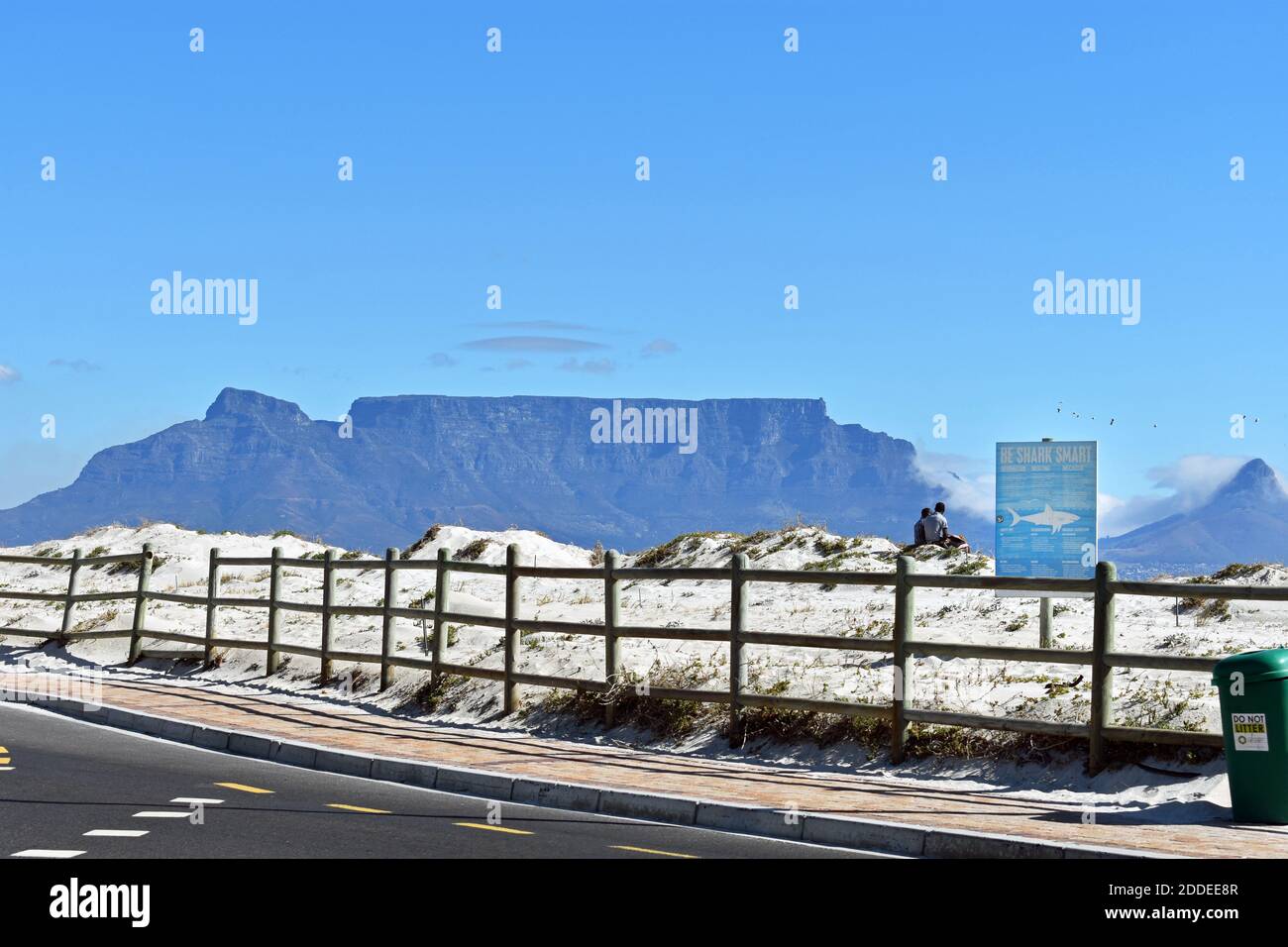 Table Mountain, Devils Peak e Lions Head visti dalla strada sulla spiaggia attraverso Table Bay. Due uomini siedono sulla sabbia accanto a un cartello intelligente di essere squalo, Foto Stock