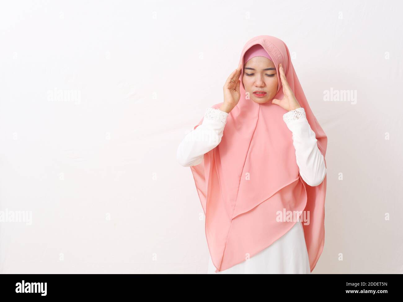 Un ritratto di donna musulmana asiatica che indossa un velo rosa o hijab e vestito bianco ha avuto un mal di testa. Toccare i templi della testa con entrambe le mani e la sofferenza Foto Stock