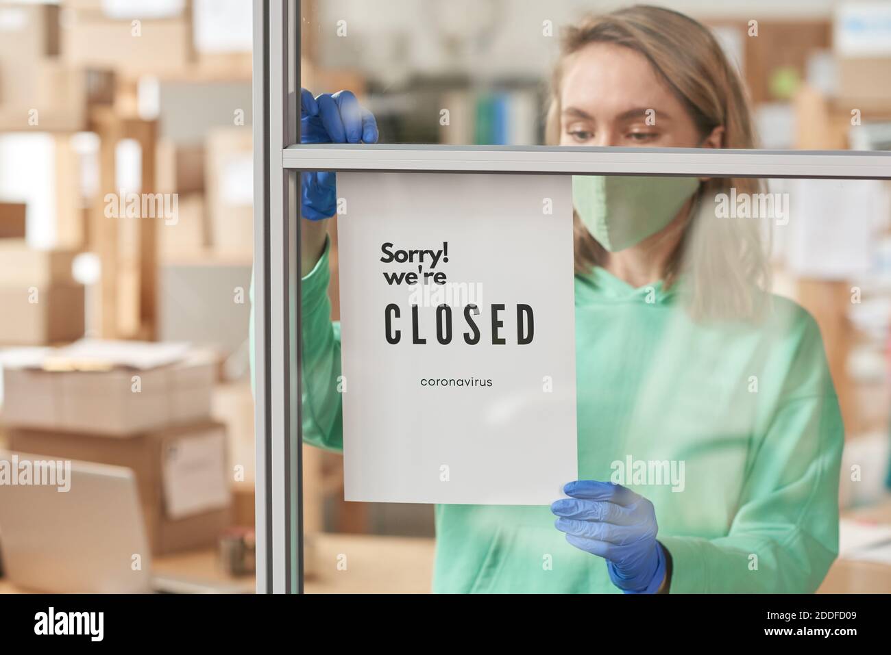 Giovane donna in maschera protettiva che appende l'annuncio sul parete di vetro con testo che l'ufficio ha chiuso Foto Stock
