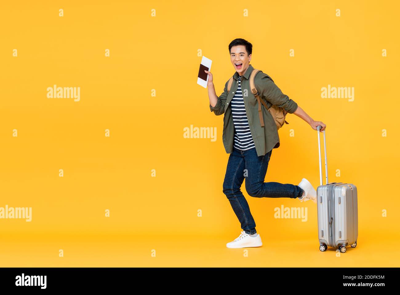 Ritratto a lunghezza intera di sorridente felice bel giovane asiatico turista con passaporto e bagaglio pronto a viaggiare in vacanza isolato in studi giallo Foto Stock