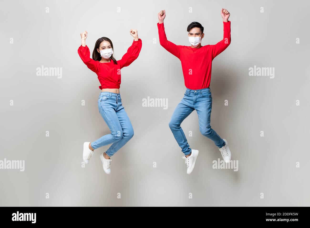 Giovane coppia asiatica indossando maschere mediche e saltando sulla luce sfondo grigio studio Foto Stock
