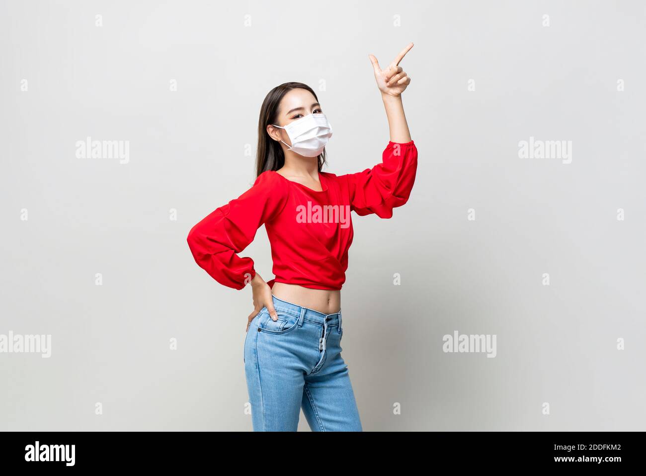Giovane donna asiatica che indossa la maschera per proteggere COVID-19 e. puntare la mano fino allo spazio vuoto isolato su grigio chiaro sfondo Foto Stock