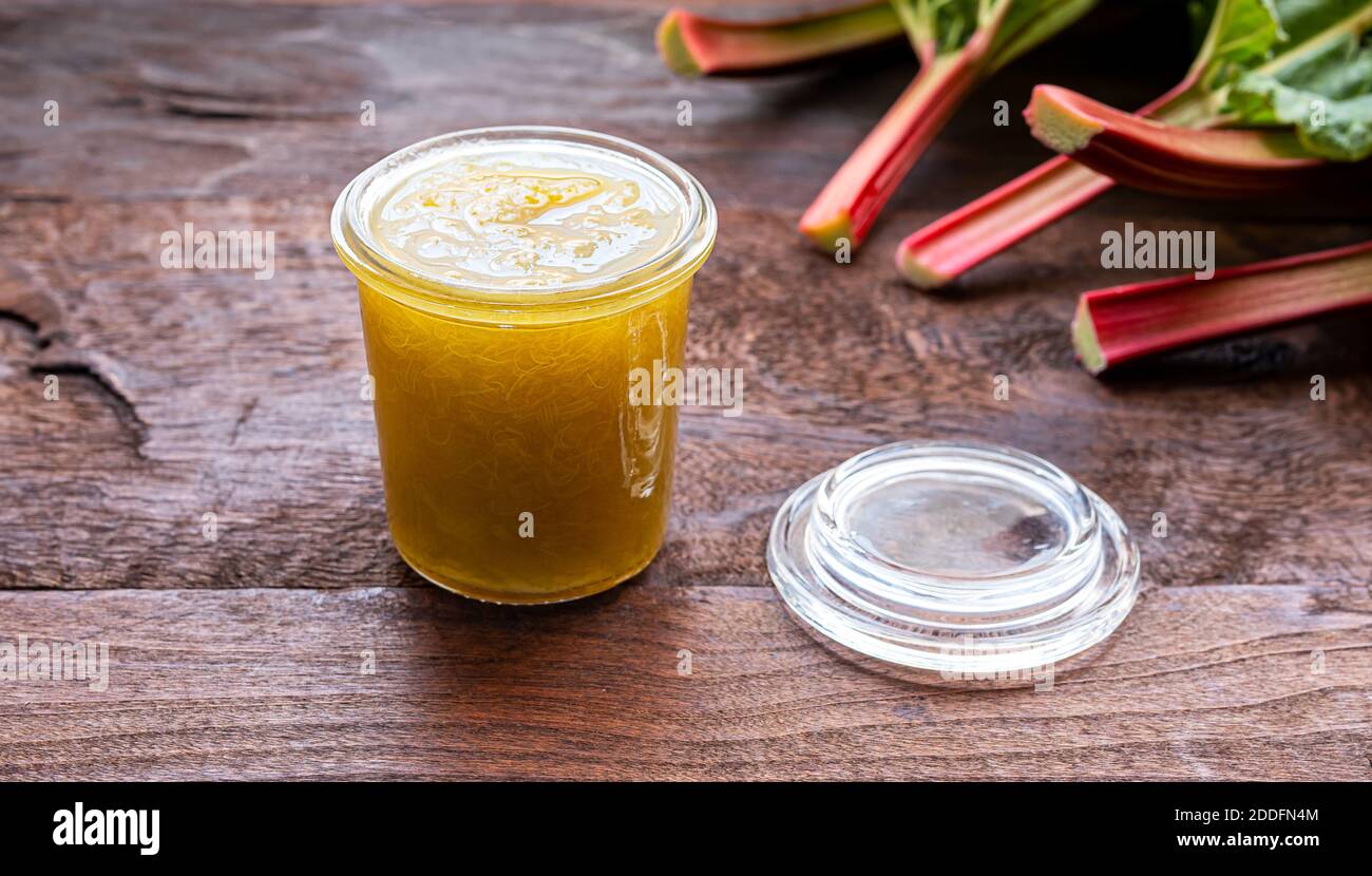 Marmellata di rabarbaro fatta in casa per una sana colazione in un vaso di vetro e gambi di rabarbaro fresco su un tavolo di legno vecchio scuro Foto Stock