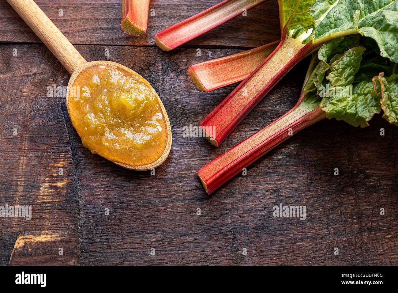 Un cucchiaio di legno con marmellata di rabarbaro fatta in casa e gambi di rabarbaro oraganico fresco su di un vecchio sfondo di legno scuro, concetto di dessert sano Foto Stock