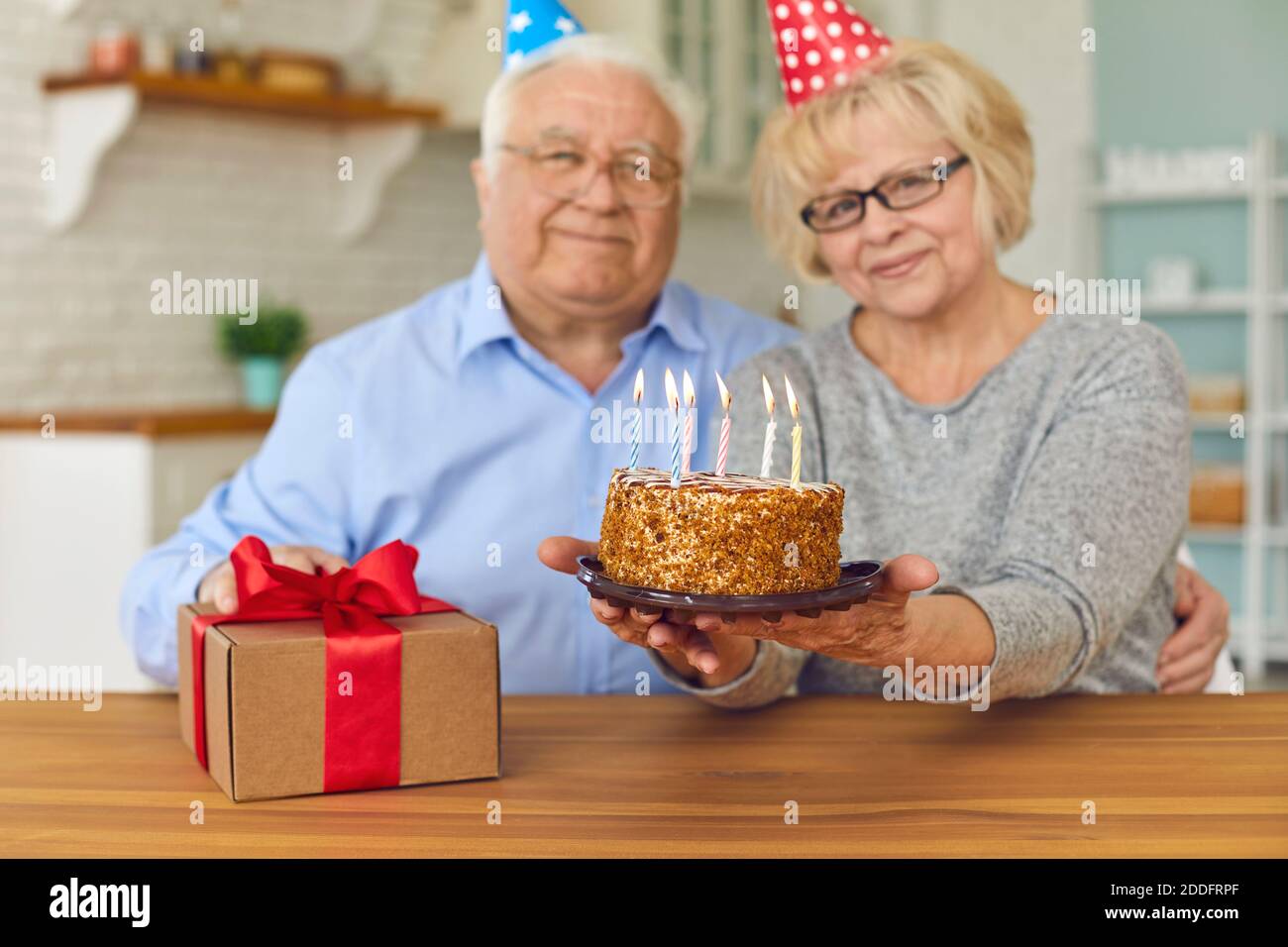 Sorridente coppia matura in caps vacanza seduta, tenendo torta di compleanno con candele e confezione regalo Foto Stock