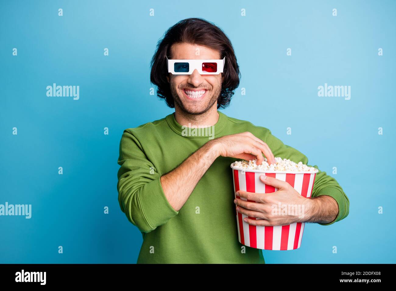 Foto di un ragazzo positivo tenere pop corn scatola mangiare sorriso lucido indossare le specifiche 3d verde pullover isolato sfondo blu Foto Stock