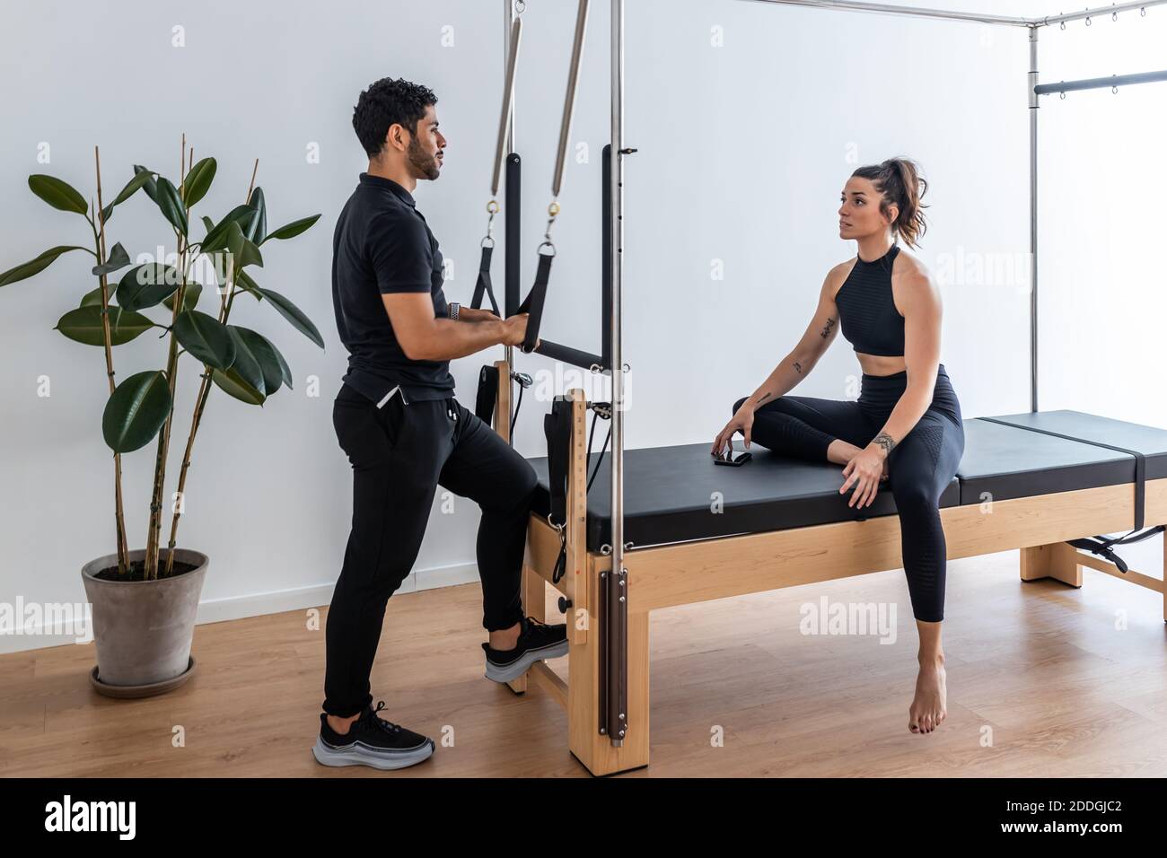 Vista laterale di uno sportivo snello seduto sul reformer cadillac pilates e parlando con un istruttore personale durante l'allenamento Foto Stock