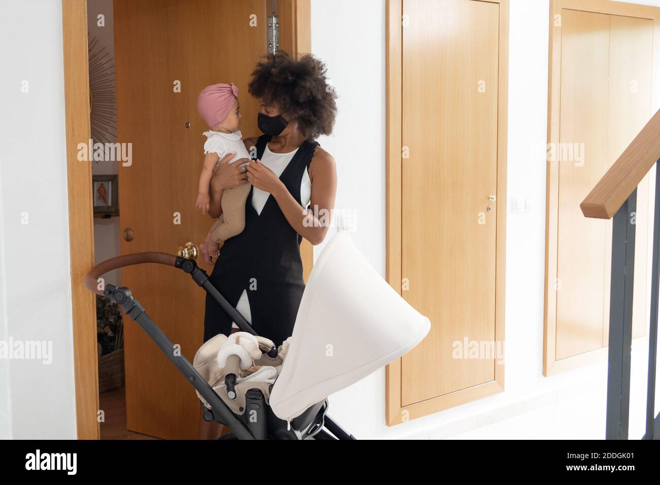 Donna Afro-americana in maschera protettiva in piedi con adorabile bambino vicino carrozza bambino mentre si prepara per passeggiare Foto Stock