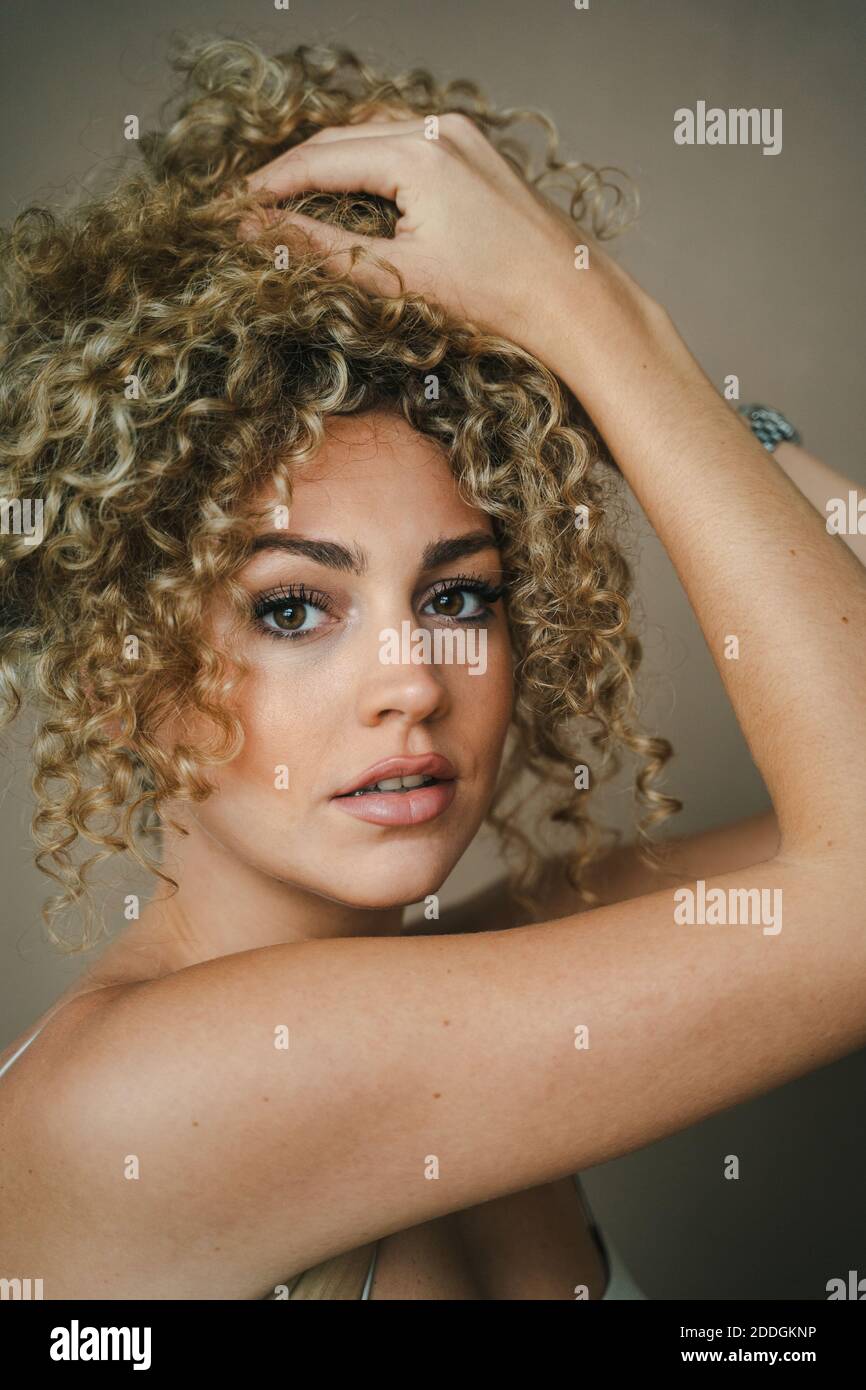 Vista laterale di affascinante donna con acconciatura e trucco Afro stare in piedi su sfondo marrone mentre si toccano i capelli e si guarda fotocamera Foto Stock