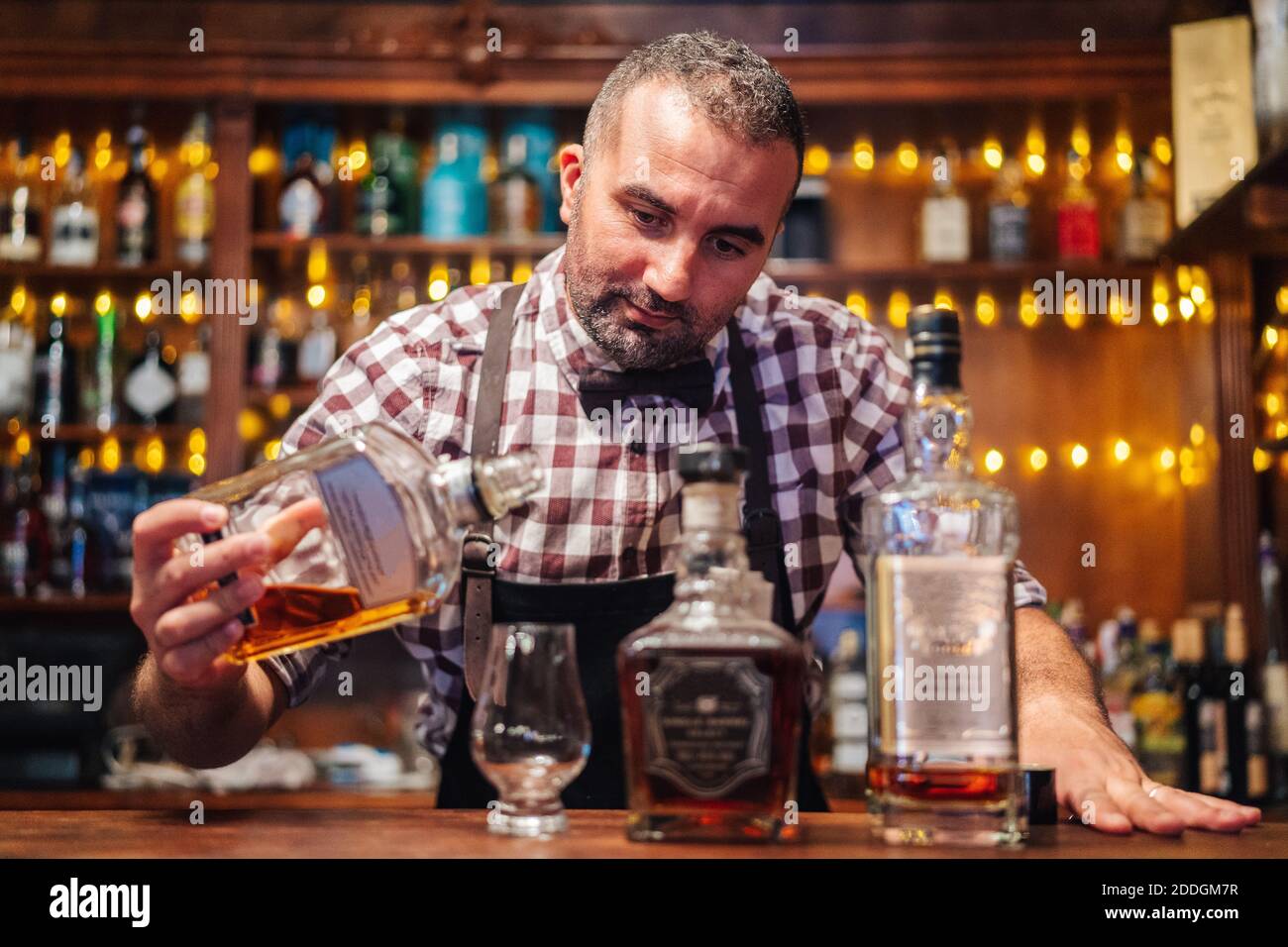 Crop brutale barista maschile in grembiule versando brandy in vetro posto sul bancone con bottiglie di bevande alcoliche Foto Stock